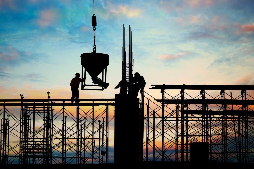 Cantiere edile al tramonto, con operai sulle impalcature e una benna sospesa alla gru